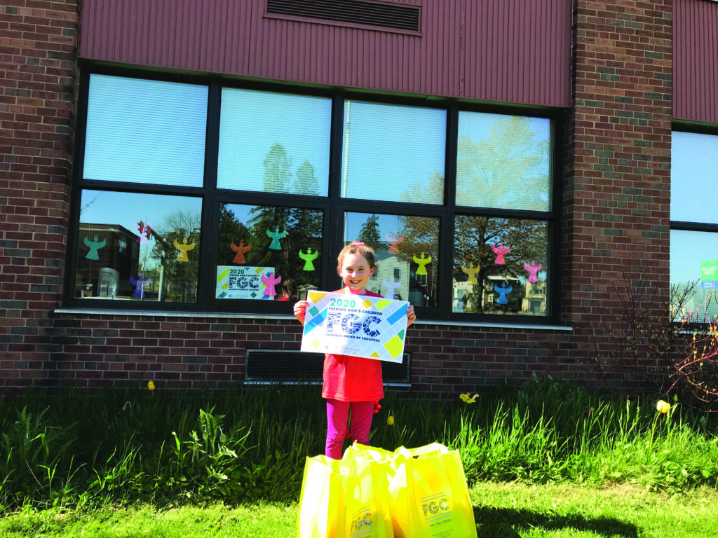 A young girl holds up a Feeding God's Children banner. She is standing in front of a building with angel cutouts in the windows. There are Feeding God's Children collection bags on the grass in front of her.