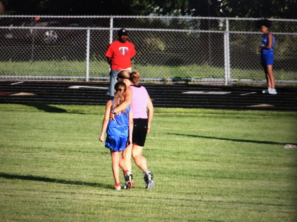 Julie has her arm around a young girl in a sports uniform and leans over to talk to the girl. They are walking on a course for track and field with their backs turned towards the camera.
