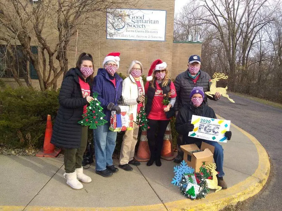 Six people are shown outside of the Good Samaritan building holding various handmade Christmas decorations such as artwork of Christmas trees, reindeer, a wreath and a gift bag. One man is holding a Feeding God's Children banner.