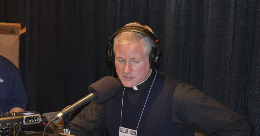 Father Rocky, a mid-aged man, is shown sitting at a table, looking at his computer and speaking into a microphone. Audio recording equipment and promotional flyers for Relevant Radio are also on the table.