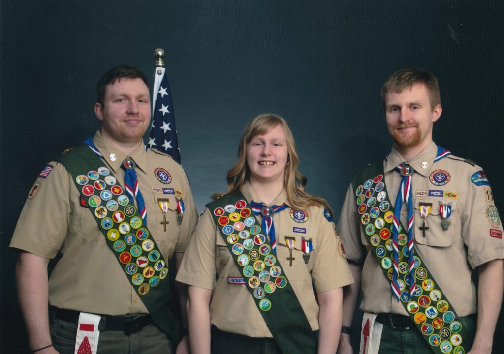 Alyxandria stands with her two brothers on either side of her. They all are wearing Eagle Scout uniforms showing their medals and many badges.