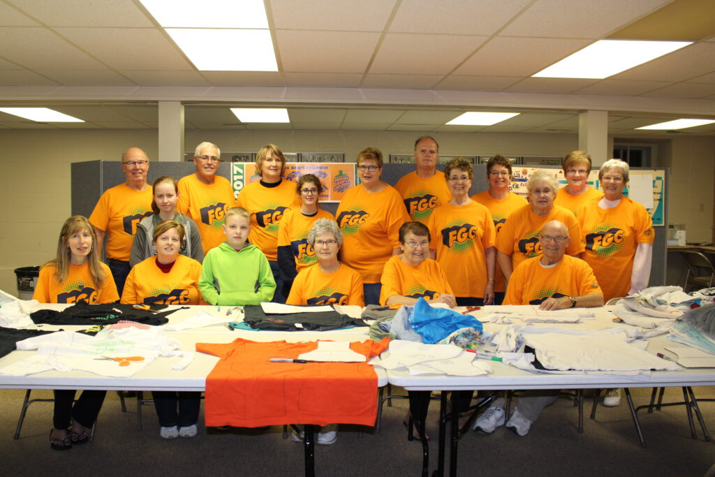 A group of volunteers in orange FGC t-shirts pose for a photo behind a table that has t-shirts, diaper patterns, and scissors on it.