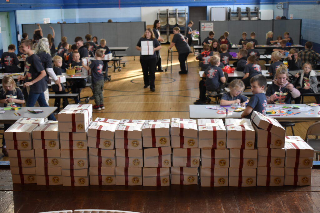 Stacked care packages are at the forefront of this image. Shown behind the boxes is a gymnasium full of student volunteers working on crafts.
