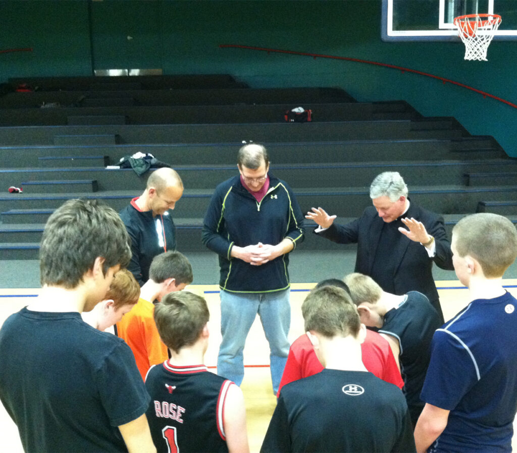 Father Rocky is shown in a gymnasium in a circle with a boy's youth basketball team. Everybody has their heads bowed, Father Rocky also has his hands outstretched in a blessing. 