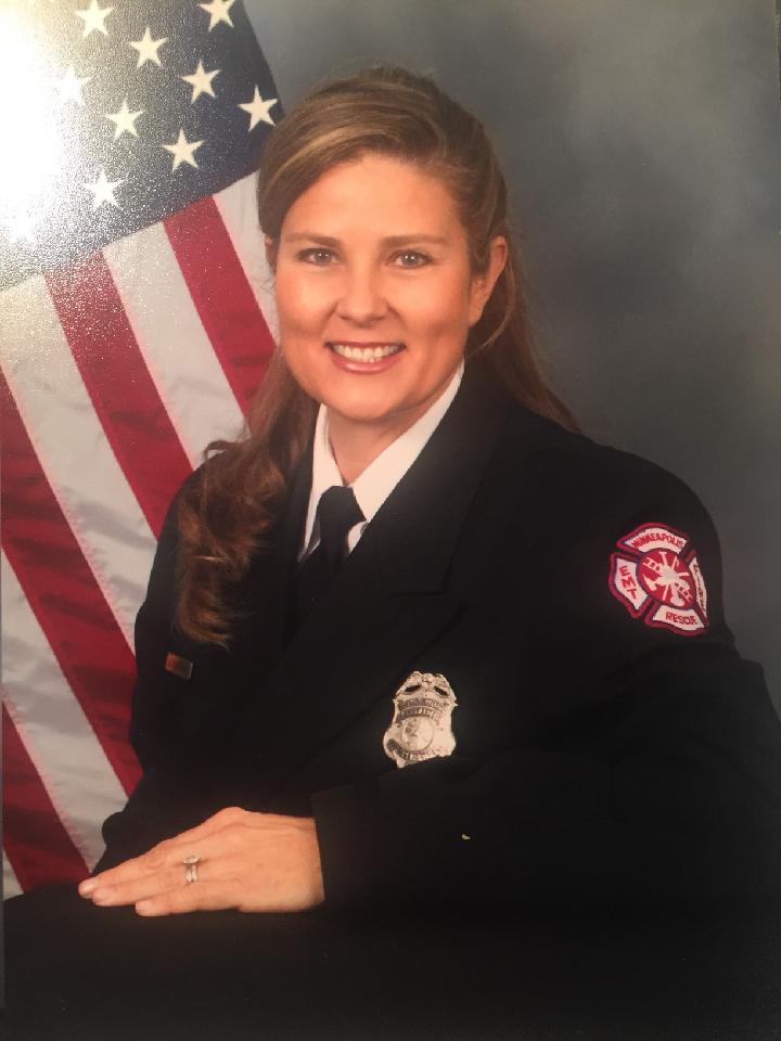 A portrait of Julie. She is wearing a uniform with her nametag, a firefighter badge, and an EMT patch that is on the shoulder. There is an American flag in the background. 
