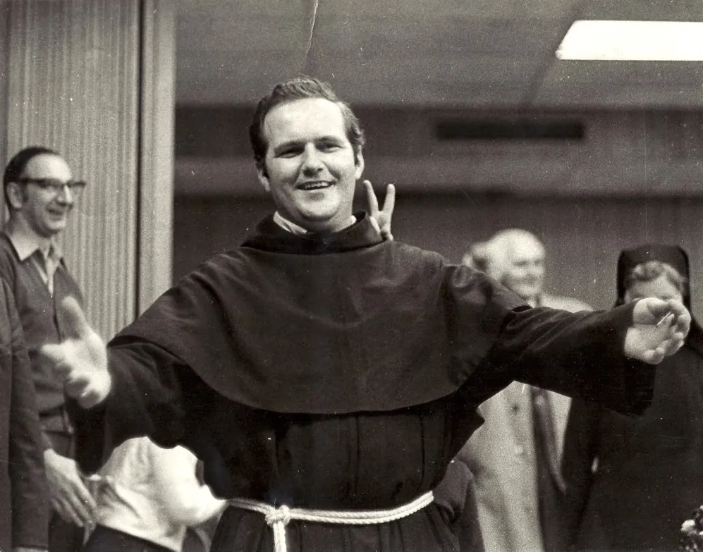 A young Father Casimir is shown wearing modest black priest robes and a rope tied around his waist. He is cheerfully smiling and has his arms outstretched. He is inside and other people can be seen behind him. 