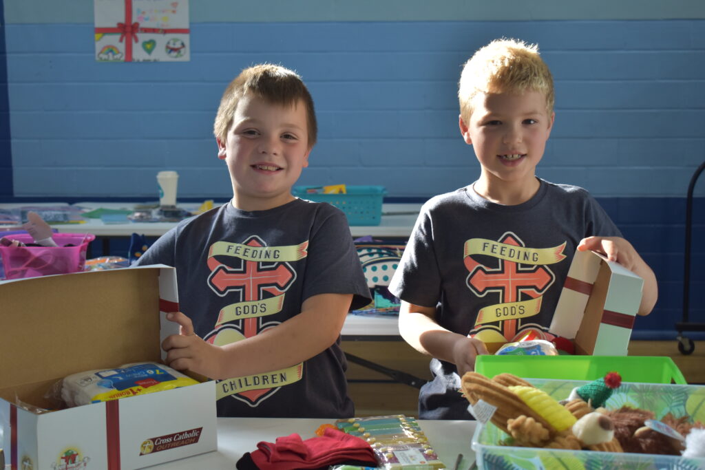 Two boys show items in the boxes they are packing, such as socks, crafts, and stuffed animals. 