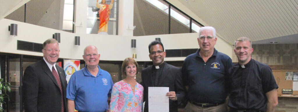 Six people stand in a church and pose for a photo, one person who is dressed like a priest holds a check.