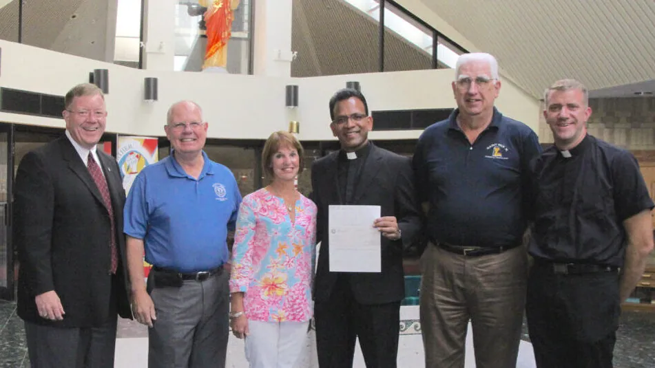 Six people stand in a church and pose for a photo, one person who is dressed like a priest holds a check.