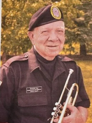 Ricardo Coy is shown wearing a uniform and holding a trumpet. 